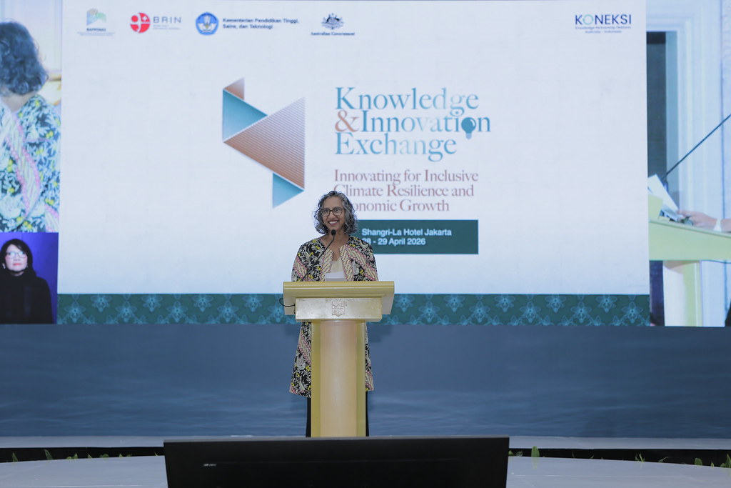 Australian Deputy Ambassador to Indonesia, Gita Kamath, delivering a speech at the podium during the Knowledge & Innovation Exchange (KIE) Jakarta Summit. The background screen displays the theme "Innovating for Inclusive Climate Resilience and Economic Growth" along with KONEKSI, Bappenas, BRIN, and Australian Government logos.